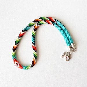 May include: A tubular beaded necklace featuring a repeating geometric pattern in vibrant turquoise, orange, green, white, and black seed beads. The necklace is finished with silver-toned metal end caps, a lobster claw clasp, and a short extender chain.