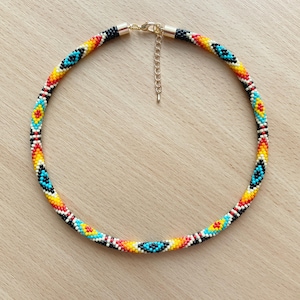 May include: A beaded necklace with a geometric pattern in black, white, red, yellow, and blue. The necklace has a gold clasp.