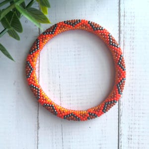 Op de afbeelding: Een ronde armband gemaakt van kleine glaskralen in een herhalend chevronpatroon. Het ontwerp bevat feloranje, donkerbruine en metallic antracietgrijze kralen die in een doorlopend zigzagmotief rondom de gehele band zijn gerangschikt.