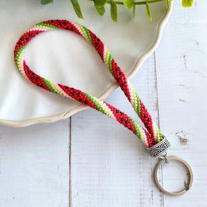 May include: A beaded lanyard in a watermelon design. The lanyard features red, green, white, and black beads, with a silver-colored metal ring and decorative clasp. The lanyard is designed to hold keys or an ID badge.