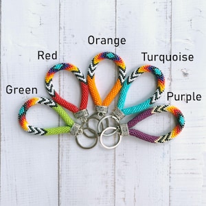 May include: Five beaded keychains arranged in a fan shape, each featuring a loop of small colorful beads with geometric patterns and a silver metal ring. The labels Green, Red, Orange, Turquoise, and Purple identify the primary color of each keychain.
