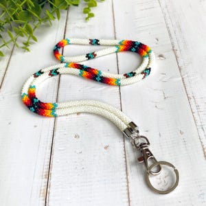 Könnte beinhalten: Ein weißes Perlen-Lanyard mit einem farbenfrohen Muster aus roten, orangefarbenen, gelben, türkisfarbenen und schwarzen Perlen. Das Lanyard hat einen silberfarbenen Verschluss und einen Schlüsselring. Der Perlenabschnitt ist ca. 91 cm lang.