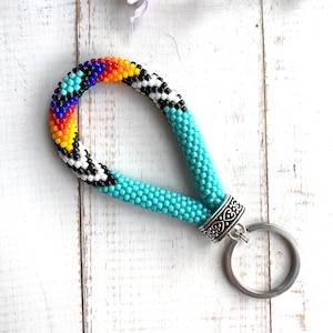 May include: A beaded keychain with a turquoise wrist strap and a silver-colored key ring. The top of the strap features a colorful pattern of red, orange, yellow, blue, and black beads. The keychain is on a white wooden surface.