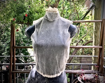 May include: A white sheer sleeveless top with a high neck and a scalloped edge. The top is on a dress form in front of a wooden fence and greenery.