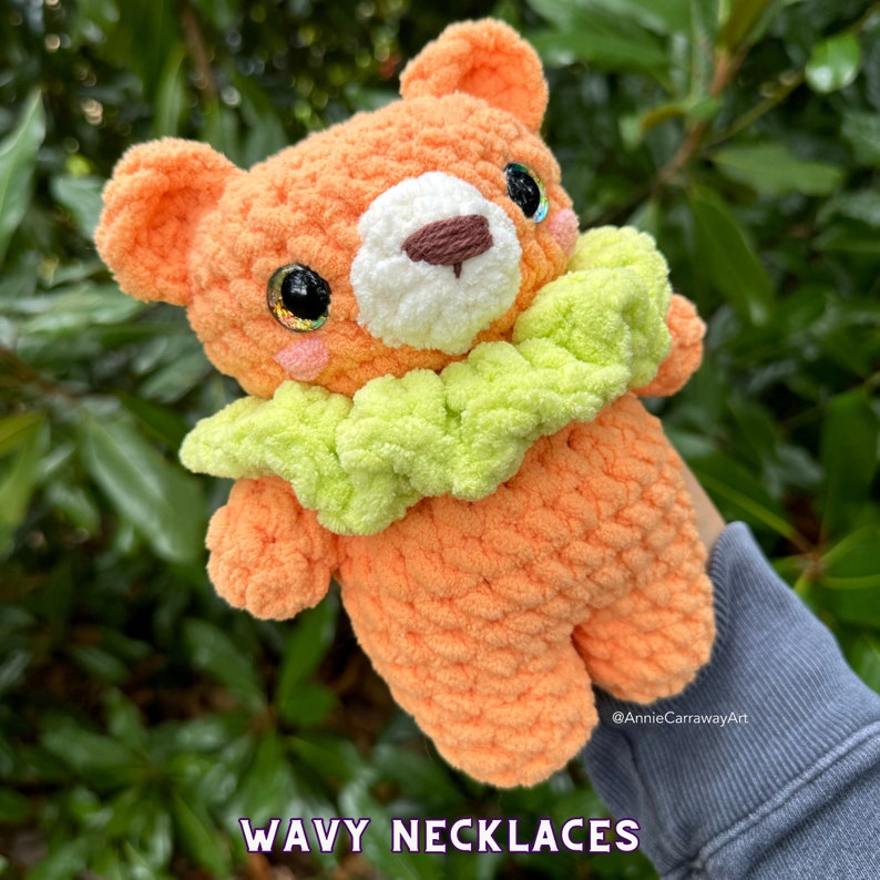 May include: A crocheted orange bear with a green and white wavy collar. The bear has sparkly green eyes and a white nose. The bear is holding a sign that says "WAVY NECKLACES".