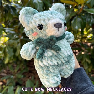 May include: A crocheted green and white teddy bear with a green bow around its neck. The bear has black eyes and a white nose. "Cute Bow Necklaces" is written in purple text at the bottom of the image.