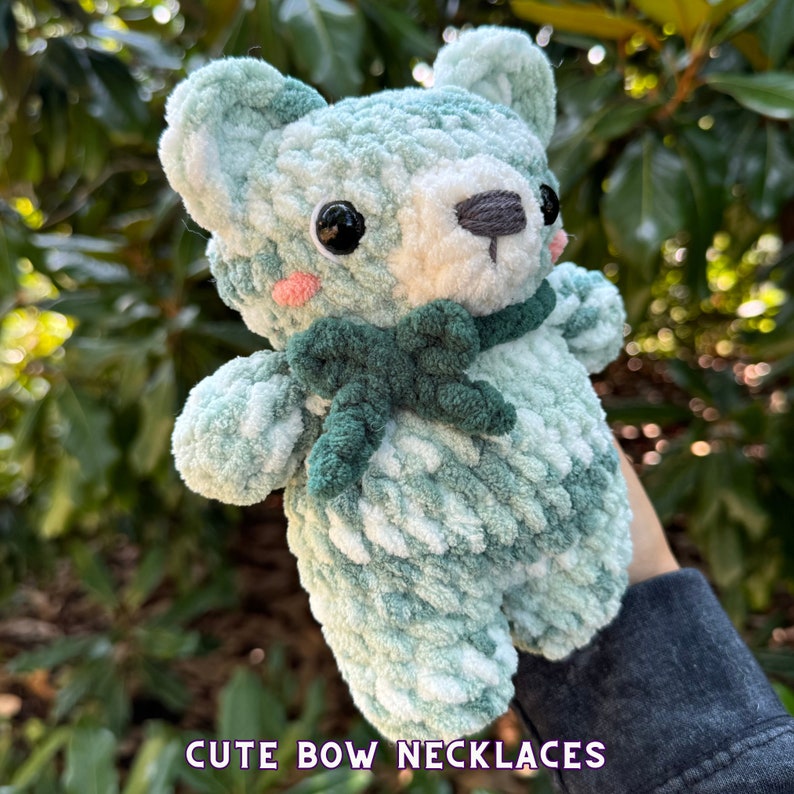 May include: A crocheted green and white teddy bear with a green bow around its neck. The bear has black eyes and a white nose. "Cute Bow Necklaces" is written in purple text at the bottom of the image.