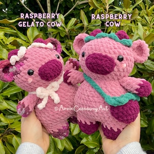 Puede incluir: Dos juguetes de peluche de crochet con forma de vaca. Uno es rosa con detalles morados y dice "Raspberry Gelato Cow". El otro es rosa con detalles verdes y dice "Raspberry Cow".