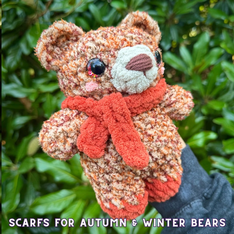 May include: A crocheted brown and orange teddy bear wearing a red scarf. The bear has large, sparkly eyes and a white nose.  The bear is holding the scarf in its paws.  Scarfs for Autumn & Winter Bears.