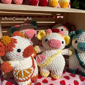 Pode incluir: Uma cole&ccedil;&atilde;o de peluches de vaca de croch&ecirc; em v&aacute;rias cores, incluindo branco, marrom, rosa e amarelo. Os peluches est&atilde;o dispostos em uma caixa de madeira com um fundo de padr&atilde;o de morango. O texto '@AnnieCarrawayArt' &eacute; vis&iacute;vel na imagem.