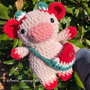 Pode incluir: Um brinquedo de pel&uacute;cia de porco rosa de croch&ecirc; com detalhes vermelhos e verdes. O porco est&aacute; segurando uma fatia de melancia na m&atilde;o direita. O porco est&aacute; usando uma al&ccedil;a listrada verde e branca.