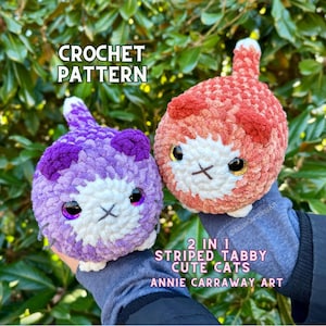 May include: Two crocheted cat amigurumi toys, one purple and one orange, with white faces and black eyes. The caption reads "2 in 1 Striped Tabby Cute Cats Annie Carraway Art".