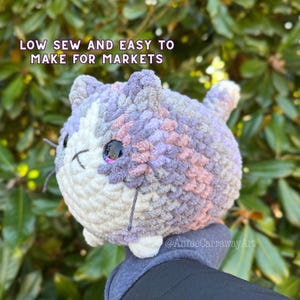 May include: A crocheted plush toy cat with a white, grey, and pink colour scheme. The cat has a large, round head and a small, pointed nose. The cat's eyes are large and black, and it has a small, pink mouth. The cat is sitting on a grey surface. The text "LOW SEW AND EASY TO MAKE FOR MARKETS" is visible above the cat.