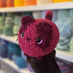 May include: A handmade, plush cat toy in shades of burgundy. The cat has large, reflective eyes, black whiskers, and a stitched 'X' on its face. The toy is round and has small ears and a tail.