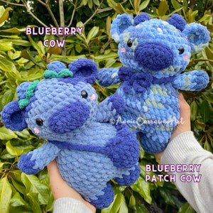 Puede incluir: Dos peluches de vaca de ar&aacute;ndano tejidos a crochet. Uno es de color azul s&oacute;lido y el otro tiene un dise&ntilde;o de patchwork con hilo azul, verde y blanco. Los peluches tienen ojos negros, mejillas rosadas y un lazo verde.