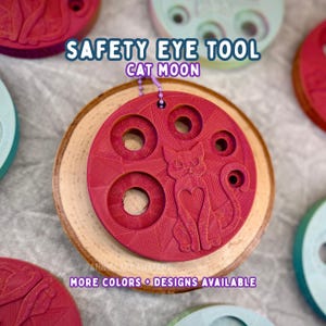 May include: A red safety eye tool with the text "SAFETY EYE TOOL CAT MOON" and a cat design. The tool has multiple circular openings and is attached to a wooden slice. The text "MORE COLORS + DESIGNS AVAILABLE" is also visible.