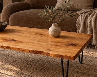 May include: A rectangular coffee table featuring a natural live-edge wood slab top with visible grain and knots, supported by four slender black metal hairpin legs. The surface displays a warm, light brown finish with a raw, organic edge profile.