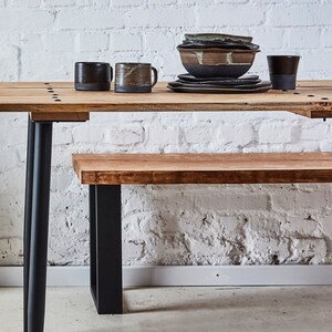 May include: A wooden table and bench set with black metal legs. The table has a natural wood finish and the bench has a light wood finish. The table is set with a bowl of ceramic dishes and two mugs.