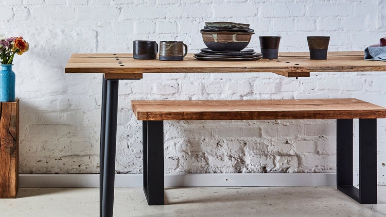May include: A wooden table and bench set with black metal legs. The table has a natural wood finish and the bench has a light wood finish. The table is set with a bowl of ceramic dishes and two mugs.