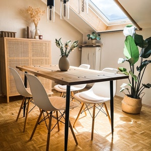 Könnte beinhalten: Ein Esstisch aus Holz mit schwarzen Metallbeinen und vier weißen Stühlen. Der Tisch ist mit einer Vase mit weißen Blumen und einer Pflanze in einem geflochtenen Korb gedeckt.