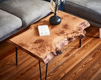 May include: A rustic wooden coffee table with a live edge top and black hairpin legs. The table is made of natural wood with a rich brown color and a unique grain pattern. A white book is placed on the table.
