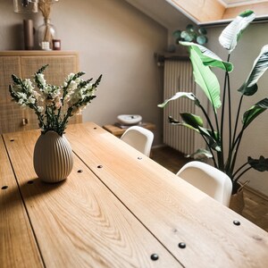 Puede incluir: Una mesa de comedor de madera con un jarr&oacute;n blanco de flores blancas en el centro. La mesa tiene detalles en metal negro y est&aacute; rodeada de sillas blancas. Una gran planta verde est&aacute; en el fondo.