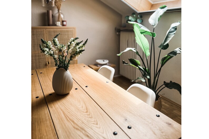 Puede incluir: Una mesa de comedor de madera con un jarr&oacute;n blanco de flores blancas en el centro. La mesa tiene detalles en metal negro y est&aacute; rodeada de sillas blancas. Una gran planta verde est&aacute; en el fondo.