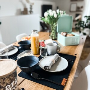 Puede incluir: Una mesa de desayuno con un mantel individual negro, un plato gris, un taz&oacute;n negro, una cuchara negra, una servilleta gris, un vaso de agua, una botella de jugo de naranja, un taz&oacute;n de fruta y una caja de pan azul claro con la palabra "Pan" en ella.