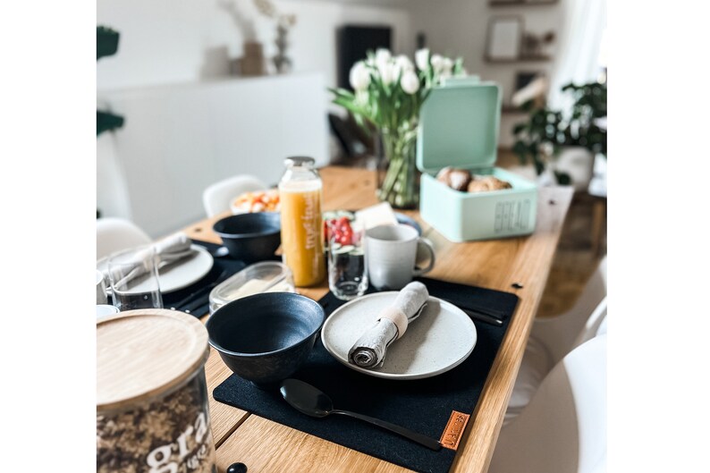 Puede incluir: Una mesa de desayuno con un mantel individual negro, un plato gris, un taz&oacute;n negro, una cuchara negra, una servilleta gris, un vaso de agua, una botella de jugo de naranja, un taz&oacute;n de fruta y una caja de pan azul claro con la palabra "Pan" en ella.