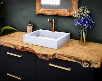 May include: A white rectangular vessel sink with a copper tap sits on a wooden countertop with a live edge. The countertop is mounted on a black cabinet with gold drawer pulls. A vase with blue flowers and a soap dispenser are on the countertop.