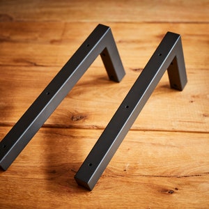 Könnte beinhalten: Zwei schwarze Metall-Regalhalterungen mit quadratischer Form und abgerundeten Kanten. Die Halterungen befinden sich auf einer Holzoberfläche.