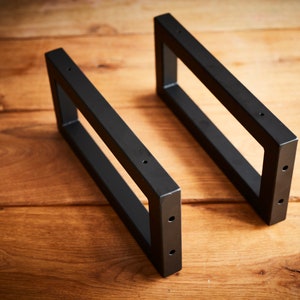 Könnte beinhalten: Zwei schwarze Metall-Regalhalterungen mit rechteckiger Form und abgerundeten Ecken. Die Halterungen haben an jeder Seite vier Löcher zum Befestigen.