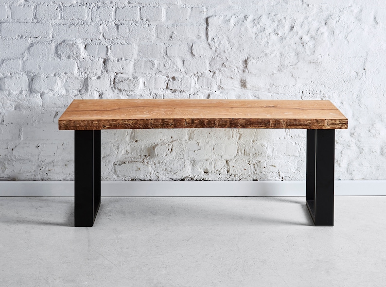 May include: A wooden table with a natural wood finish and black metal legs. The table is sitting on a concrete floor in front of a white brick wall.