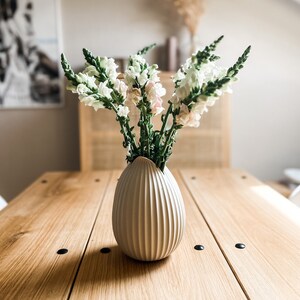 Puede incluir: Un jarr&oacute;n de cer&aacute;mica blanco con textura acanalada, que contiene un ramo de flores de boca de drag&oacute;n blancas y rosas. El jarr&oacute;n est&aacute; sobre una mesa de madera.