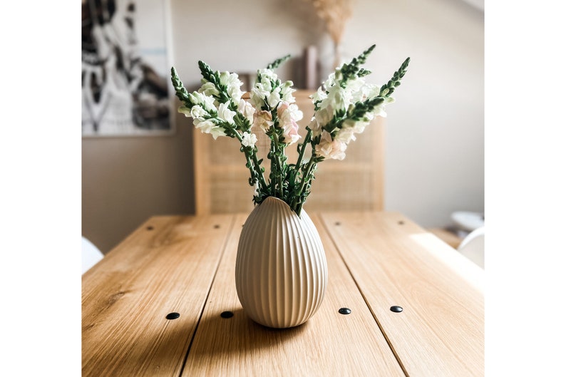 Puede incluir: Un jarr&oacute;n de cer&aacute;mica blanco con textura acanalada, que contiene un ramo de flores de boca de drag&oacute;n blancas y rosas. El jarr&oacute;n est&aacute; sobre una mesa de madera.