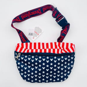 Puede incluir: Ri&ntilde;onera azul marino con cabezas de Mickey Mouse y estrellas blancas. La parte superior tiene rayas rojas y blancas. La correa ajustable es roja con "Disneyland Resort" impreso en blanco. Hebilla azul.