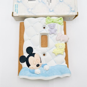 May include: A decorative light switch plate featuring Mickey Mouse sleeping on a blue quilted bed. The plate has a white background with a green bow, a purple teddy bear, a pink bunny, and a yellow sheep.