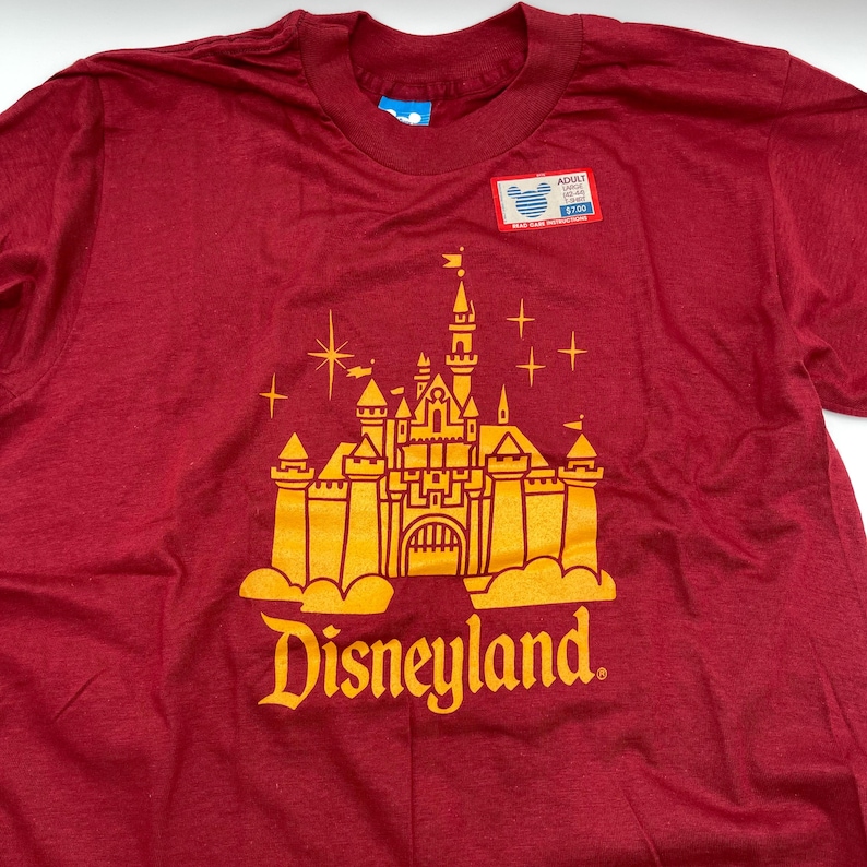 May include: A maroon short-sleeve t-shirt featuring a yellow graphic of a castle with stars above it and the text Disneyland below. A rectangular tag is attached to the chest area with the text ADULT LARGE 42-44 L-RED.