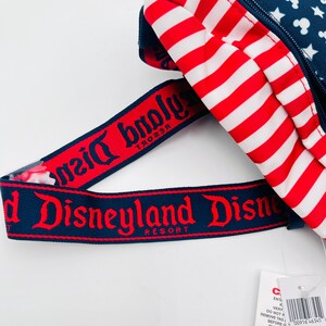 Puede incluir: Una mochila roja, blanca y azul con un patr&oacute;n de rayas y una correa azul marino. La correa presenta las palabras "Disneyland Resort" en letras rojas. La mochila tiene una secci&oacute;n azul marino con estrellas blancas y siluetas de Mickey Mouse.