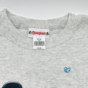 May include: A light gray heathered t-shirt featuring a white neck label with the red Disneyland logo and two small Mickey Mouse icons. A white retail tag with a barcode and a small Mickey Mouse graphic is attached to the neck label.