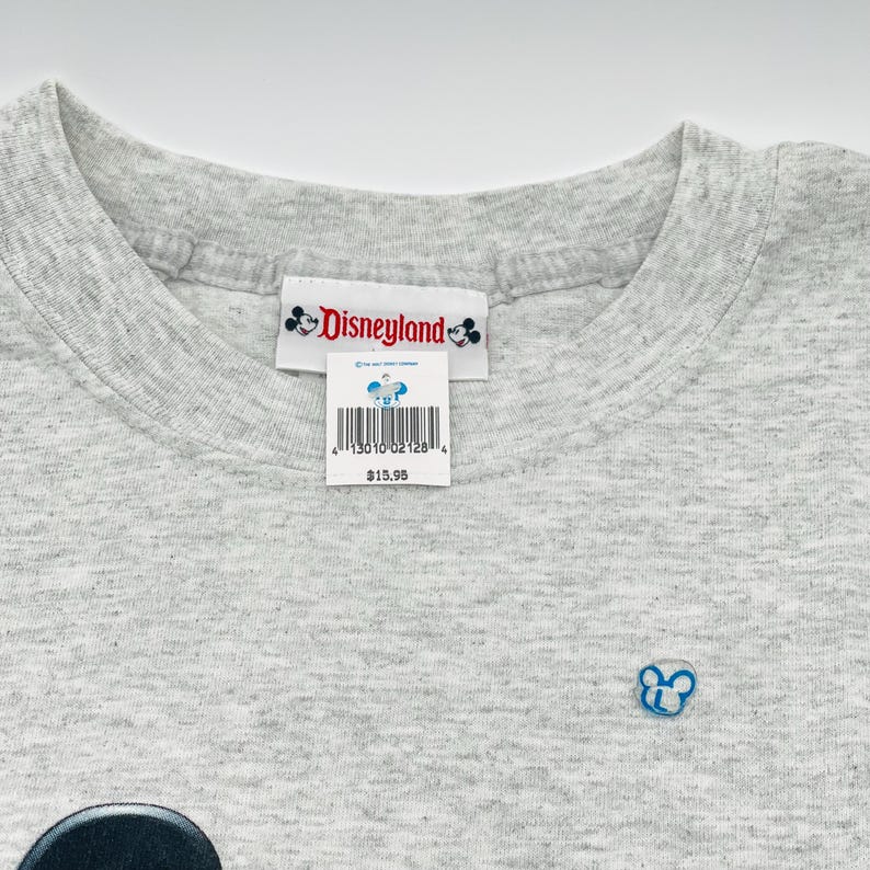May include: A light gray heathered t-shirt featuring a white neck label with the red Disneyland logo and two small Mickey Mouse icons. A white retail tag with a barcode and a small Mickey Mouse graphic is attached to the neck label.