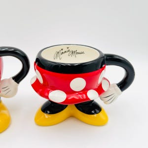 Op de afbeelding: Een Minnie Mouse-thema mok met een zwart handvat en rand, een rode body met witte stippen en gele voeten. De binnenkant is wit met "Minnie Mouse" in cursief. De mok is ontworpen om op Minnie Mouse te lijken.