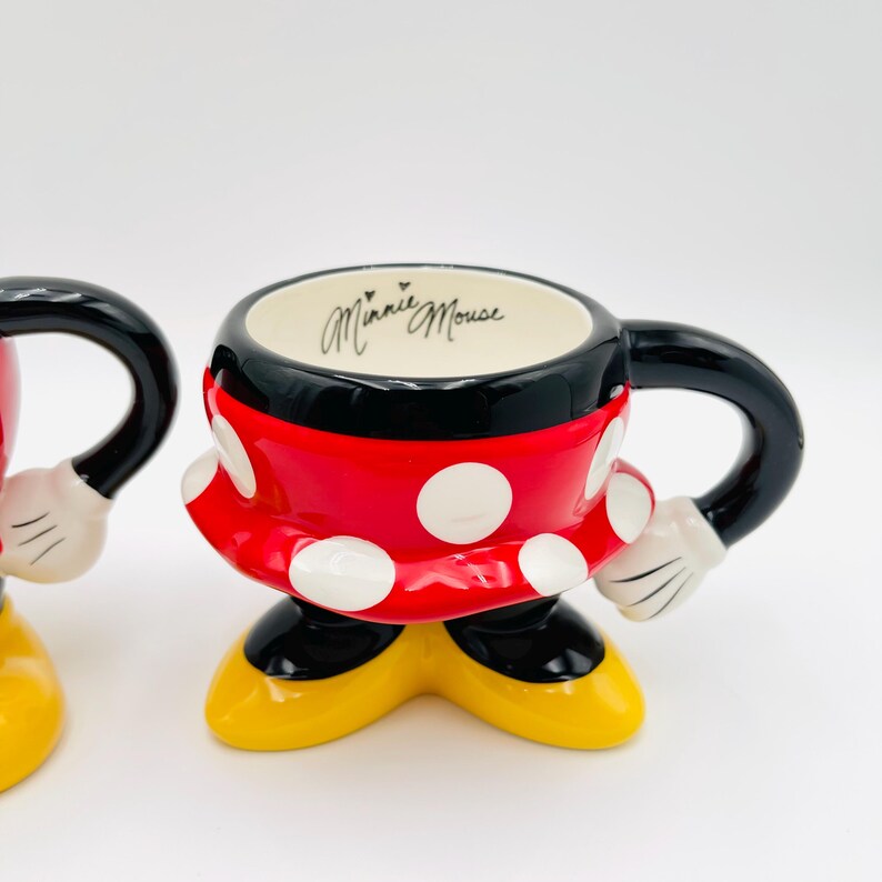 Op de afbeelding: Een Minnie Mouse-thema mok met een zwart handvat en rand, een rode body met witte stippen en gele voeten. De binnenkant is wit met "Minnie Mouse" in cursief. De mok is ontworpen om op Minnie Mouse te lijken.
