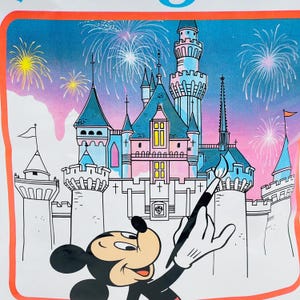 May include: A graphic print featuring Mickey Mouse holding a paintbrush in front of a large castle with blue and pink towers. The background displays a night sky filled with colorful fireworks, all enclosed within a bright orange rectangular border.