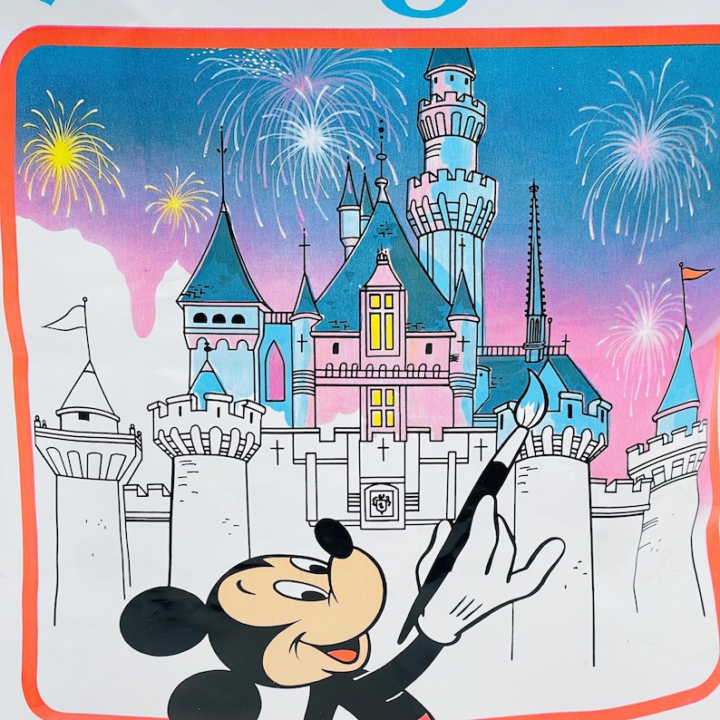 May include: A graphic print featuring Mickey Mouse holding a paintbrush in front of a large castle with blue and pink towers. The background displays a night sky filled with colorful fireworks, all enclosed within a bright orange rectangular border.