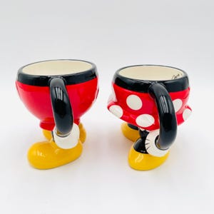 Op de afbeelding: Twee keramische mokken, &eacute;&eacute;n rood met een zwart handvat en de andere rood met witte stippen, in de vorm van Mickey en Minnie Mouse. De mokken hebben gele voeten en witte handschoenen. De mokken zijn ontworpen om eruit te zien als de iconische outfits van de personages.