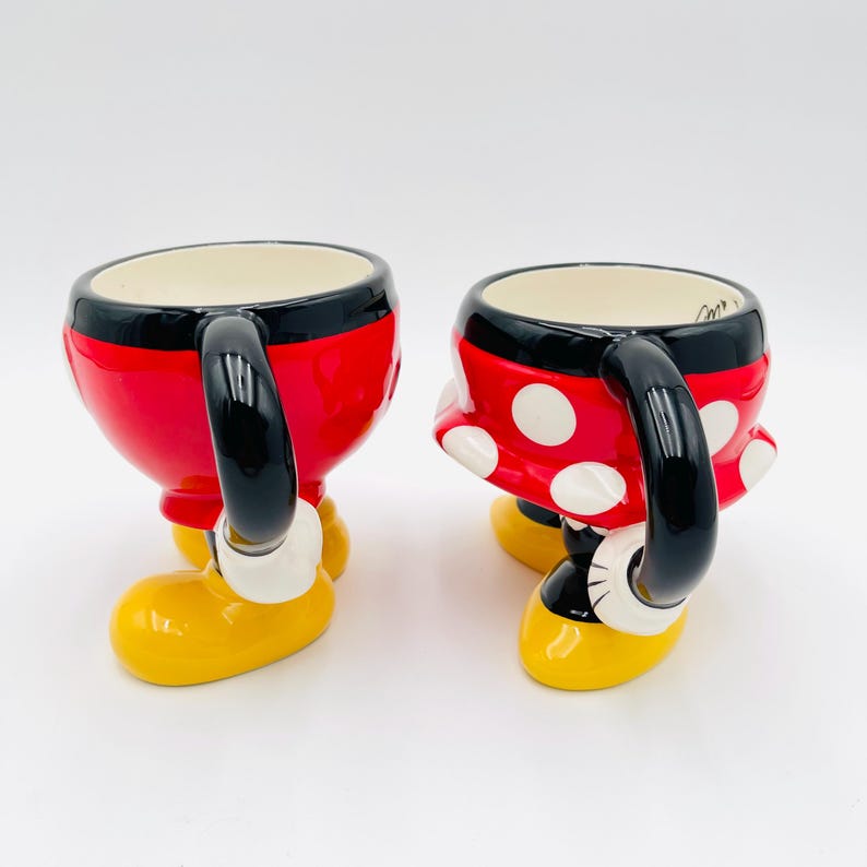 Op de afbeelding: Twee keramische mokken, &eacute;&eacute;n rood met een zwart handvat en de andere rood met witte stippen, in de vorm van Mickey en Minnie Mouse. De mokken hebben gele voeten en witte handschoenen. De mokken zijn ontworpen om eruit te zien als de iconische outfits van de personages.