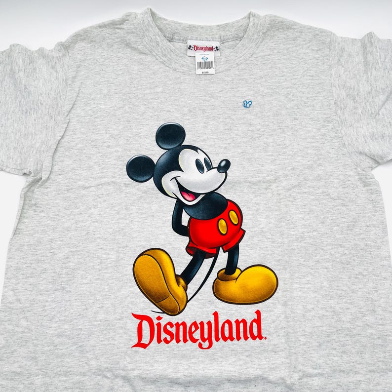 May include: A heather gray short-sleeve t-shirt featuring a graphic of Mickey Mouse wearing red shorts and yellow shoes. Below the character is the red text "Disneyland". The inner neck label displays the "Disneyland" logo and a barcode.