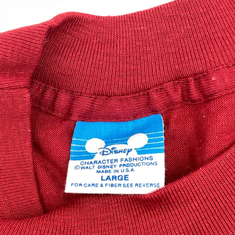 May include: A red t-shirt featuring a blue and white rectangular tag at the neckline. The tag displays the Disney logo, the text CHARACTER FASHIONS, WALT DISNEY PRODUCTIONS, MADE IN U.S.A., LARGE, and FOR CARE & FIBER SEE REVERSE.