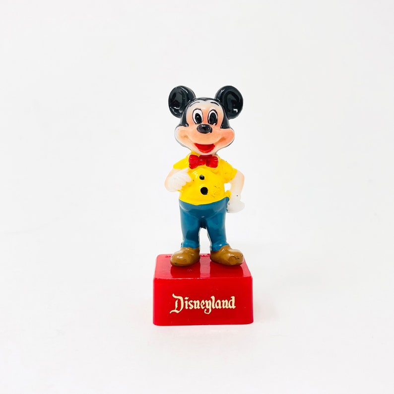 May include: A Mickey Mouse figurine, wearing a yellow shirt, blue trousers, and a red bow tie, stands on a red base. The base is inscribed with the word "Disneyland" in gold lettering. The figurine has black ears and brown shoes.